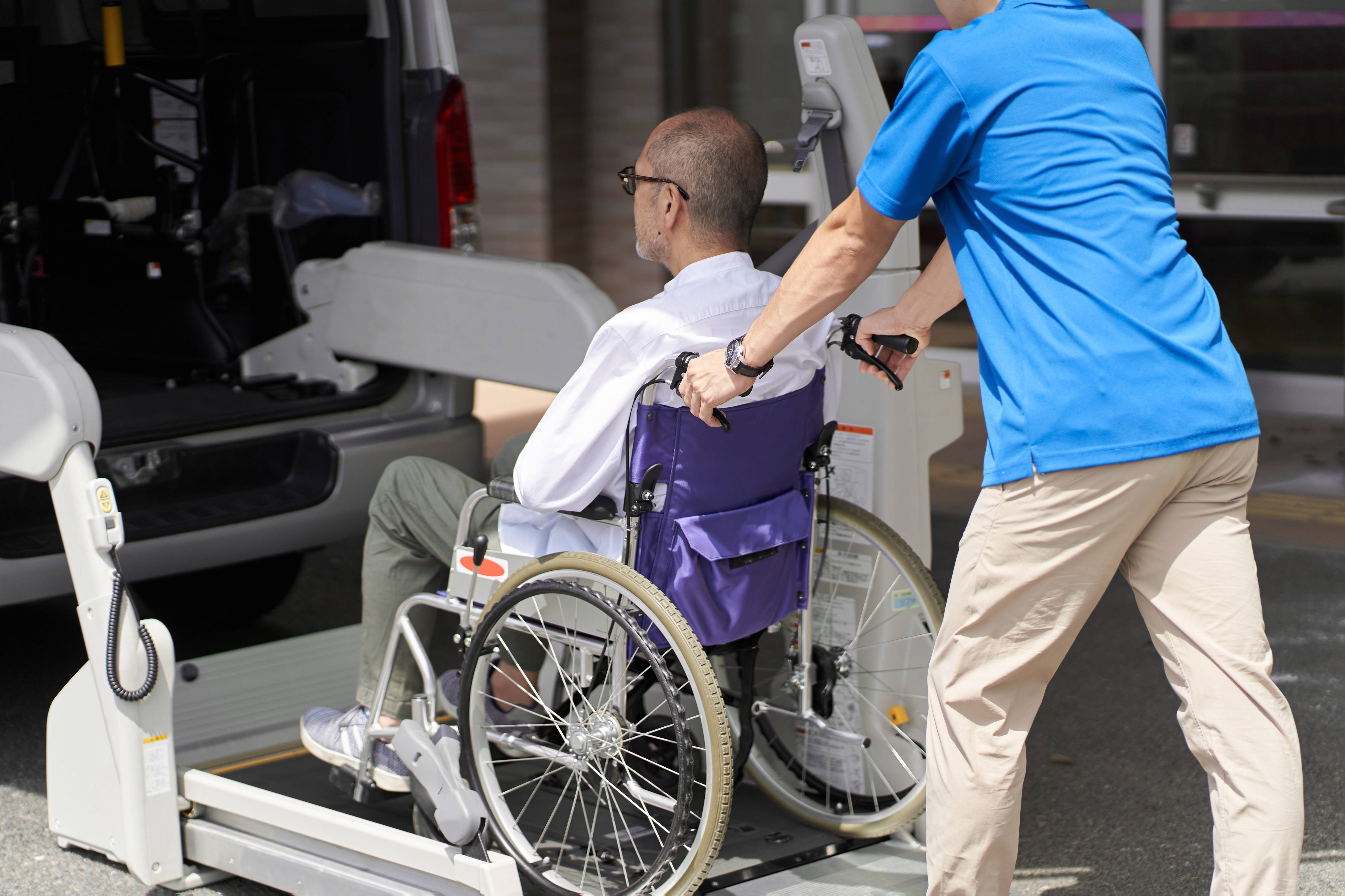 Professional JA Care Transport staff member in blue uniform carefully assisting an elderly gentleman in wheelchair onto the vehicle's hydraulic wheelchair lift, demonstrating our safe and compassionate medical transportation services